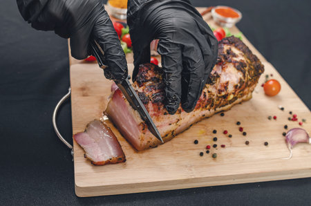 Slicing pork belly with gloved hands using knife. Close-up of gloved hands slicing delicious piece of roasted pork belly on wooden cutting board with pepper and cherry tomatoes, on black background.の写真素材