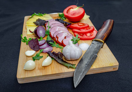Knife for slicing. Slices of meat and cheese on wooden board with fresh garlic cloves, sliced tomato, onion and pepper. Black background.の写真素材