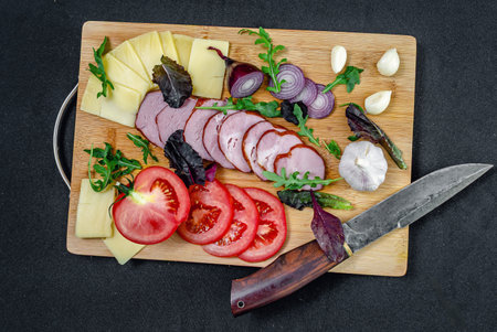 Delicious slices of baked pork ham and cheese on wooden board with fresh garlic cloves, sliced tomato, blue onion and colored pepper. Knife for slicing. Top view.の写真素材
