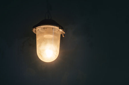 Antique street lamp on iron chain with plastic shade that shines dim yellow light at night. Worn wall background.の写真素材