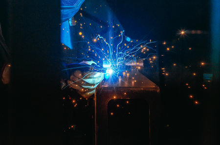 Man in protective suit welds metal part in workshop. Sparks, light and smoke in poorly lit...