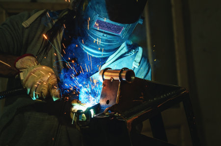 Man in protective suit welds metal part in workshop. Sparks, light and smoke in poorly lit...