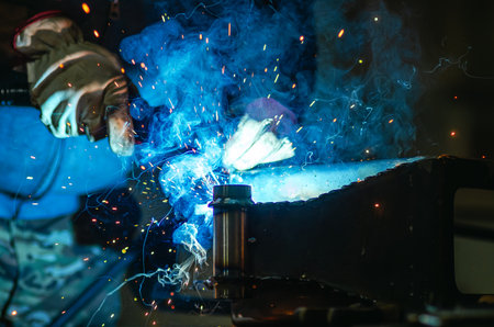 Metal welder works welding steel parts at factory in protective gloves. Production of metal structures and repair and construction services. Smoke, sparks.の写真素材