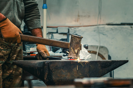 Hands of craftsman in gloves with iron hammer align iron part on stone. Vintage construction tool. Preparation for welding work.の写真素材