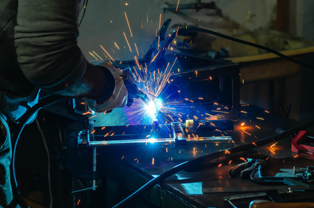 Male worker working with metal product and welding it with welding machine in workshop. Small...