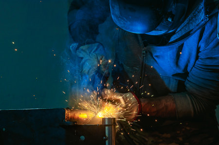 Welding shop worker in protective helmet and gloves at workplace on blue background. Blue light of...