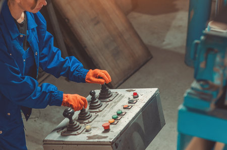 Industrial worker in blue protective uniform and orange gloves operating control panel with...