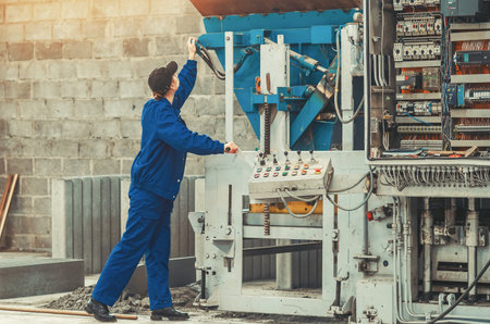 Industrial worker in blue coveralls operating heavy manufacturing equipment using control panel...