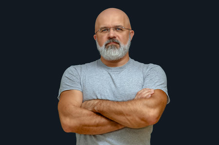 Confident mature bald man with glasses in gray t-shirt with crossed arms on dark background....