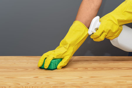 Close-up of hands wearing protective cleaning gloves wiping wooden surface with cloth and spray...