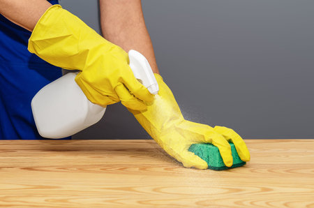 Close-up of hands wearing protective cleaning gloves wiping wooden surface with cloth and spray...