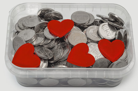 Several small red hearts placed on pile of metal coins in transparent container on neutral gray background. Concept of charity, donation, love and financial support.の写真素材