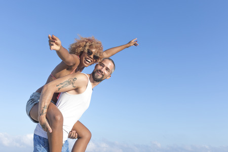 Guy carries his girlfriend piggy back style on the beach in the summer. They are laughing and smiling.の写真素材