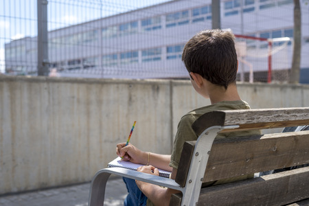 Portrait of a teenager boy sitting on a wood park bench doing his college homework and studying...