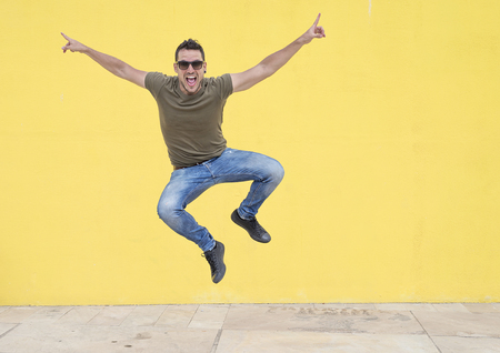 Young man with sunglasses jumping in front of a yellow wall