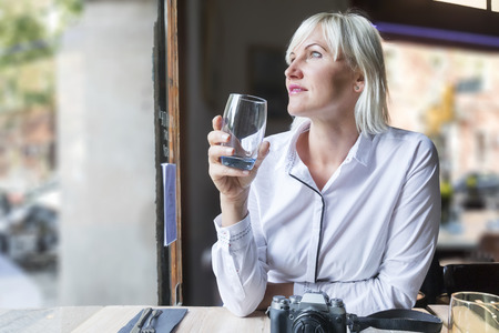 Blonde hair woman holding a glass of water next to a window of a coffeehouseの写真素材