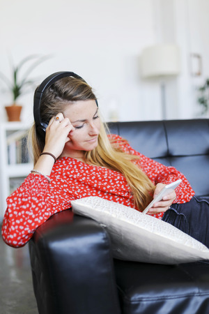 Happy woman listening to music wearing headphones using a smartphone sitting on a sofa in the living room of a interior houseの写真素材
