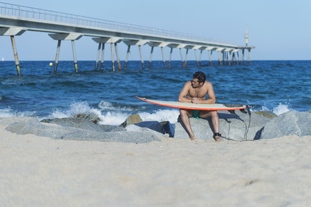 Surfer sitting on a stone, waxing his surfboard on the beach.