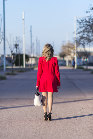 Rear image of a blonde woman wearing red jacket and holding a white handbag while walking in the cityの写真素材