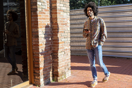 Front view of a trendy young smiling afro man using a mobile phone while standing outdoors in a sunny dayの写真素材