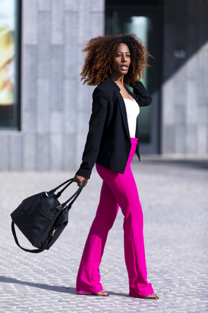 Front view of a young beautiful curly woman wearing elegant clothes and handbag while standing in the street in sunny dayの写真素材