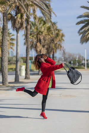 Side view of young beautiful curly afro woman wearing sunglasses smiling and dancing while standing on city path in a sunny dayの写真素材