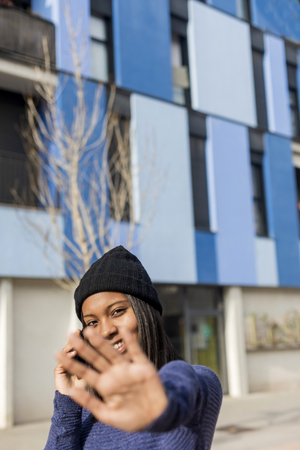 Young beautiful black woman standing outdoors in the city street using a phone while looking camera and forbidsの写真素材