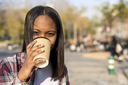 Smiling young african american woman standing in city street while holding a take away coffee and looking camera in a sunny dayの写真素材