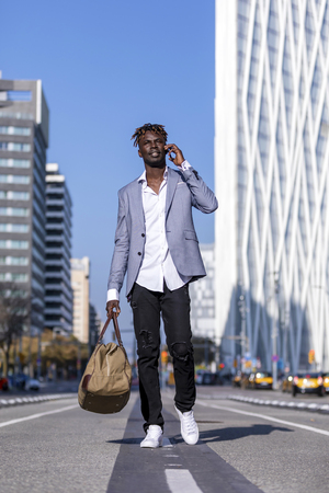 Front view of black african young man walking on the street wearing elegant jacket and holding a bag while using phone in sunny dayの写真素材