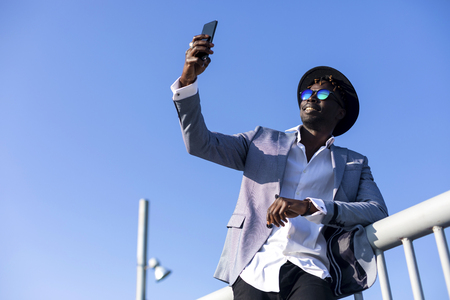 low angle view of a young african black man wearing hat and sunglasses leaning on a metallic fence relaxing while taking a selfie in a sunny day against blue skyの写真素材