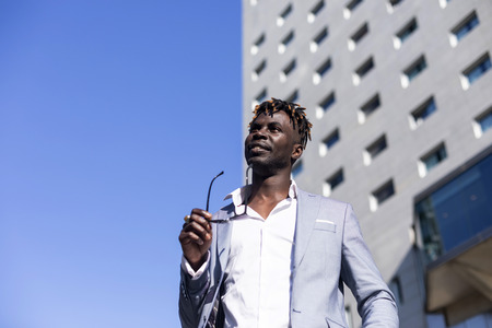 Low angle view of a black african young man wearing elegant clothes with sunglasses put in on eyeglasses while walking on the street in a sunny dayの写真素材