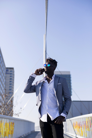 Front view of a smiling young black african man wearing elegant clothes standing in the street while using a mobile phone outdoors in sunny dayの写真素材