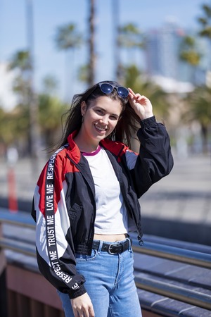 Front view of a beautiful smiling young woman wearing urban clothes and sunglasses standing while looking camera outdoors in the street in a bright dayの写真素材