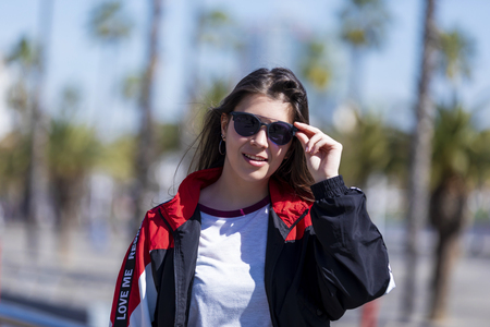 Front view of a beautiful smiling young woman wearing urban clothes and sunglasses standing while looking camera outdoors in the street in a bright dayの写真素材
