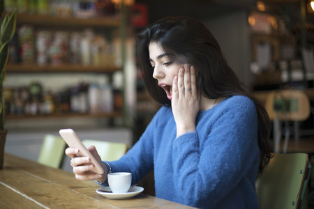 Side view of a casual girl watching smartphone and looking amazed with news while sitting in cafeteriaの写真素材