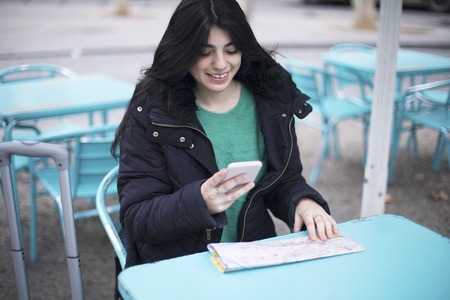 Young beautiful woman tourist sitting in a cafe outdoors, using smartphone, holding destination map.