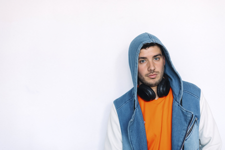 young handsome man wearing jeans jacket with hood on and headphones on his neck standing against white wallの写真素材