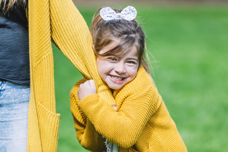 Front view of smiling little girl with a head tie standing in the park and looking at camera while holding hands with mom in a sunny dayの写真素材