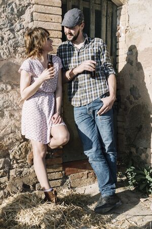 Young hipster couple in love leaning on a rural wall holding beer while bonding.の写真素材