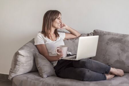 Beautiful woman relaxing on the sofa holding a cup of coffee while using laptop