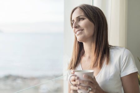 Woman standing while looking through window tasting coffee