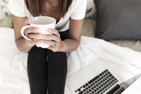 Woman sitting on bed while holding a cup of tea, working with laptop