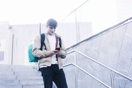 Young man walking down stairs with backpack while using mobile outdoorsの写真素材