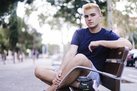 Portrait of a young teenage boy with blonde bangs sitting on a bench in the street while looking at the cameraの写真素材