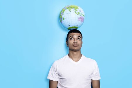 Young black male standing against blue wall while holding earth globe with his head and looking upの写真素材