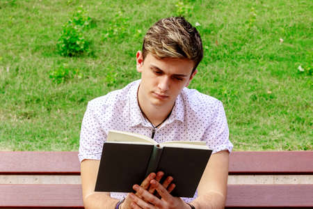 Young male reading a book while sitting on a bench in a park