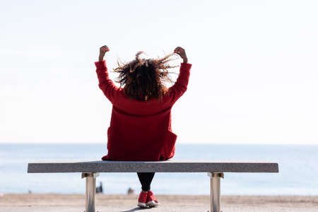 Rear view of young curly woman wearing red denim jacket sitting on a bench while looking away to...