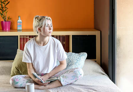 Thoughtful blond girl dressed in pajama holding a book while looking away by the windowの写真素材