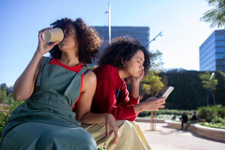 Young woman drinking coffee, sitting on bench in the street while her woman friend chatting on the...