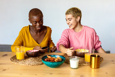 Two young women couple, best friends sitting at home, eating breakfast and drinking coffee, using smartphone.の写真素材